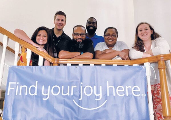A group of Sonida Senior Living team members gathered on a staircase and smiling behind a find your joy here banner.