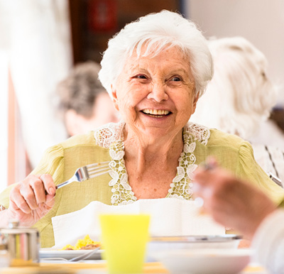 A smiling resident enjoying a cheerful, sunny meal with neighbors at a Sonida Senior Living community.