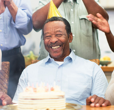 A smiling senior man in a yellow party hat celebrates his birthday with a cake and friends singing to him.