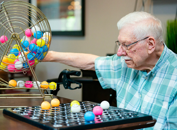 A male resident spinning a wheel and leading a group in a game of bingo at a senior living community.