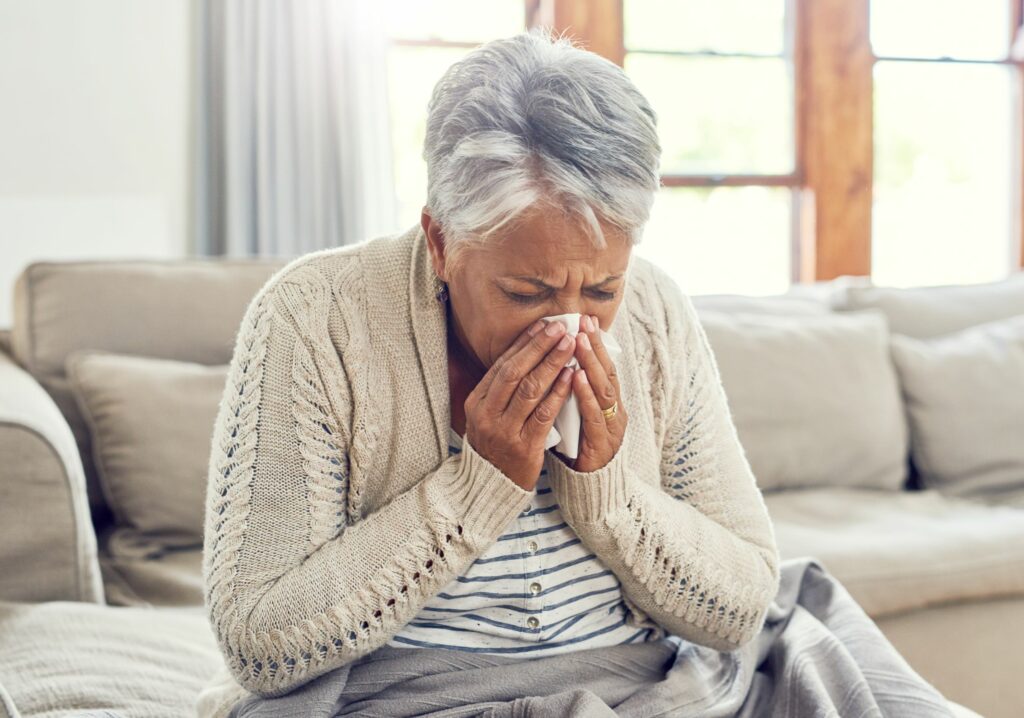 Sickly senior woman blowing her nose with a tissue while sitting on a sofa
