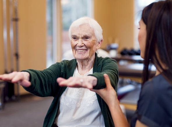 A smiling, female resident stretching her arms in front of her while assisted by a caring physical therapist in a fitness room at a Sonida Senior Living community