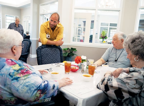 A Sonida Senior Living employee leaning on a chair and smiling as residents share a meal together in the dining hall of an independent and assisted living community.