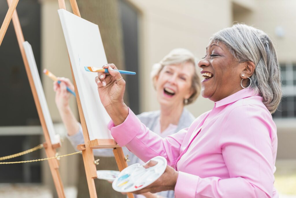 Two senior women sitting outdoors at easels painting pictures on canvases