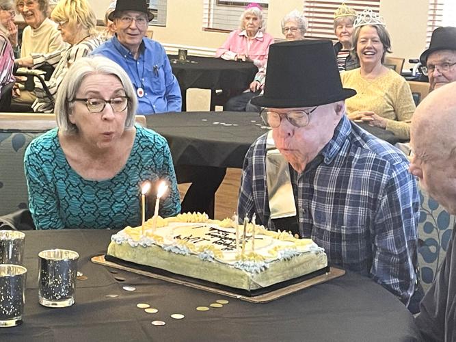 senior man blows out the candles on his birthday cake with the help of his daughter-in-law