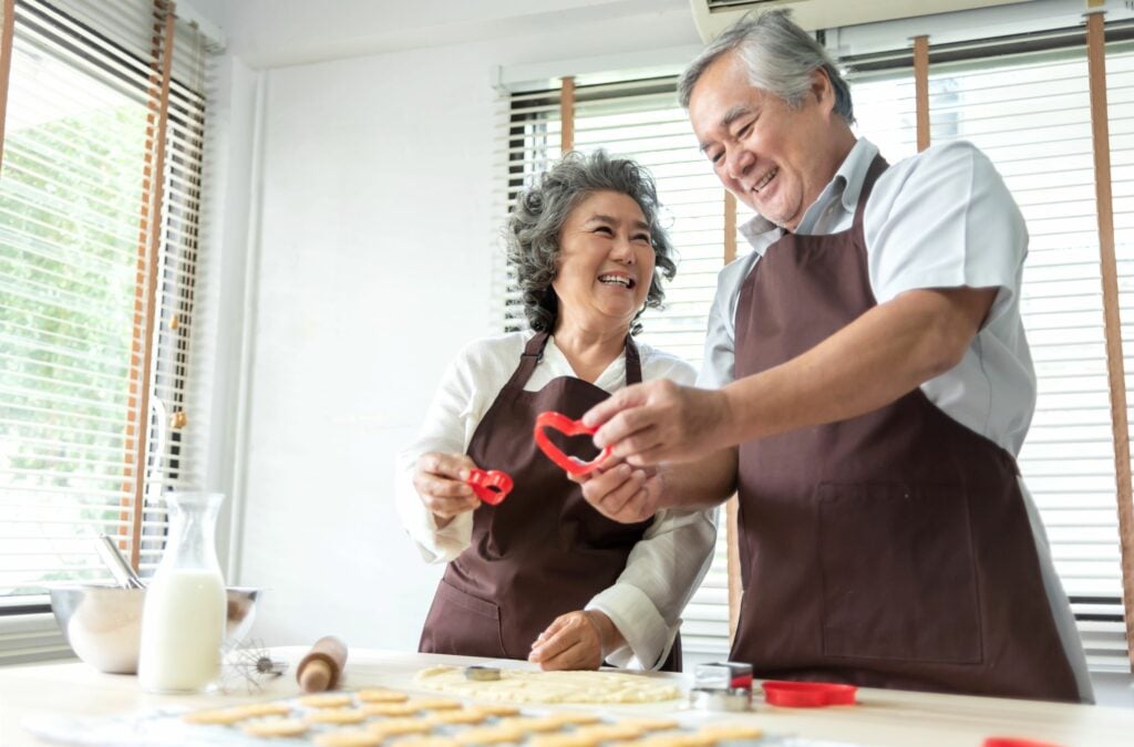 Old couple enjoying holding red cookie cutter in heart shape while preparing and kneading dough at home together