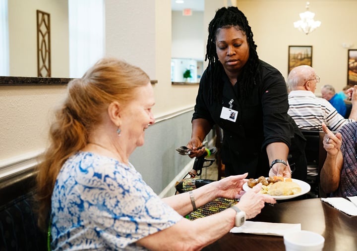 An older adult woman smiles as a plate of food is handed to her by a female senior living employee