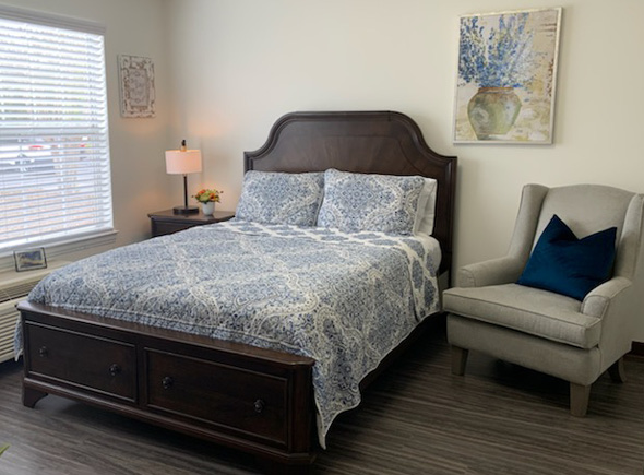 Memory care apartment bedroom with a large bed, chair, side table and window