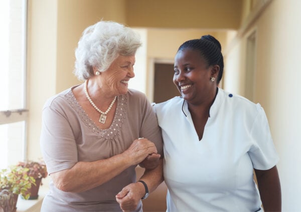 Senior woman being led down a hallway by a smiling female caregiver.