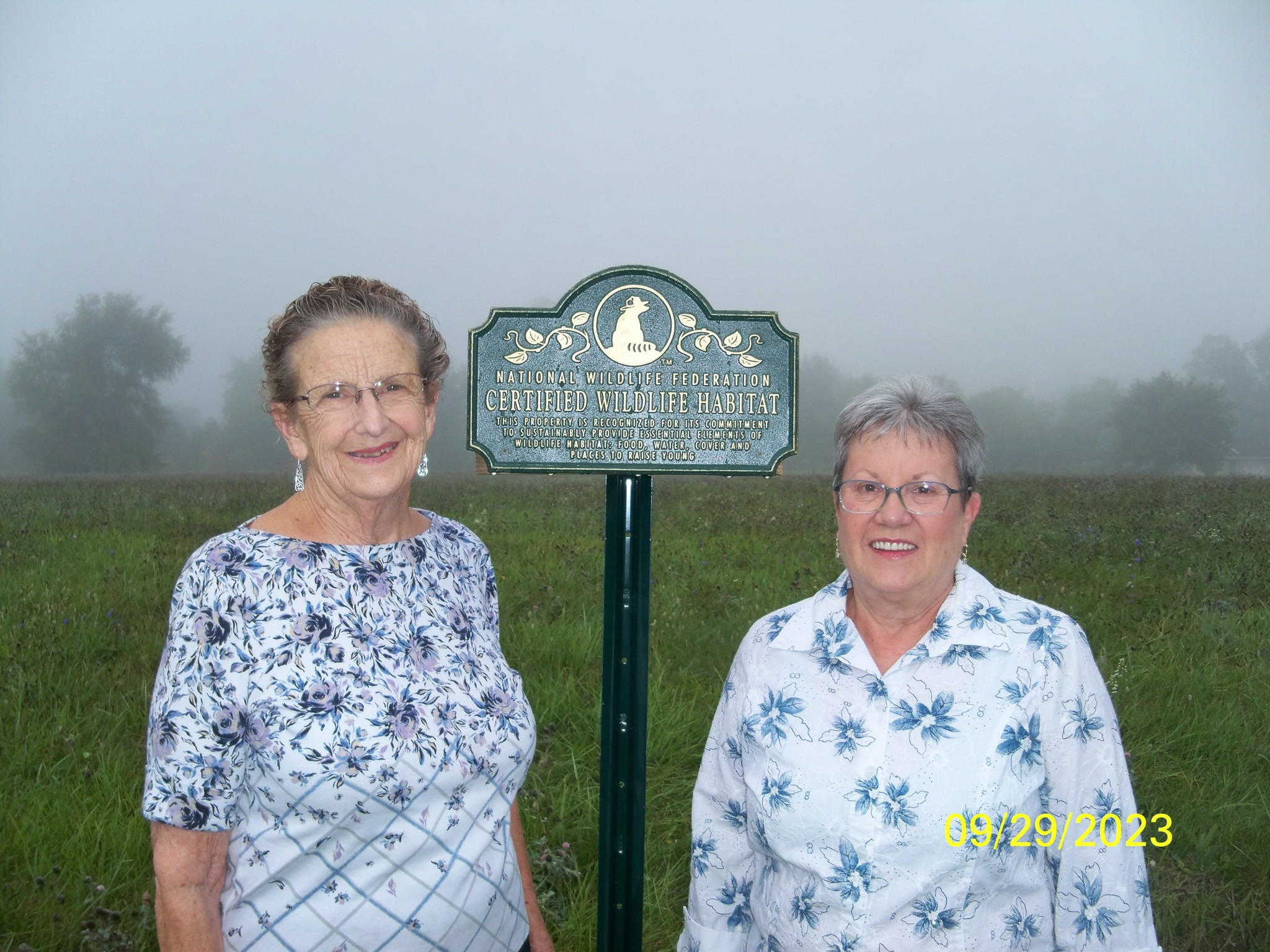 Two senior women stand in front of a sign that reads, 