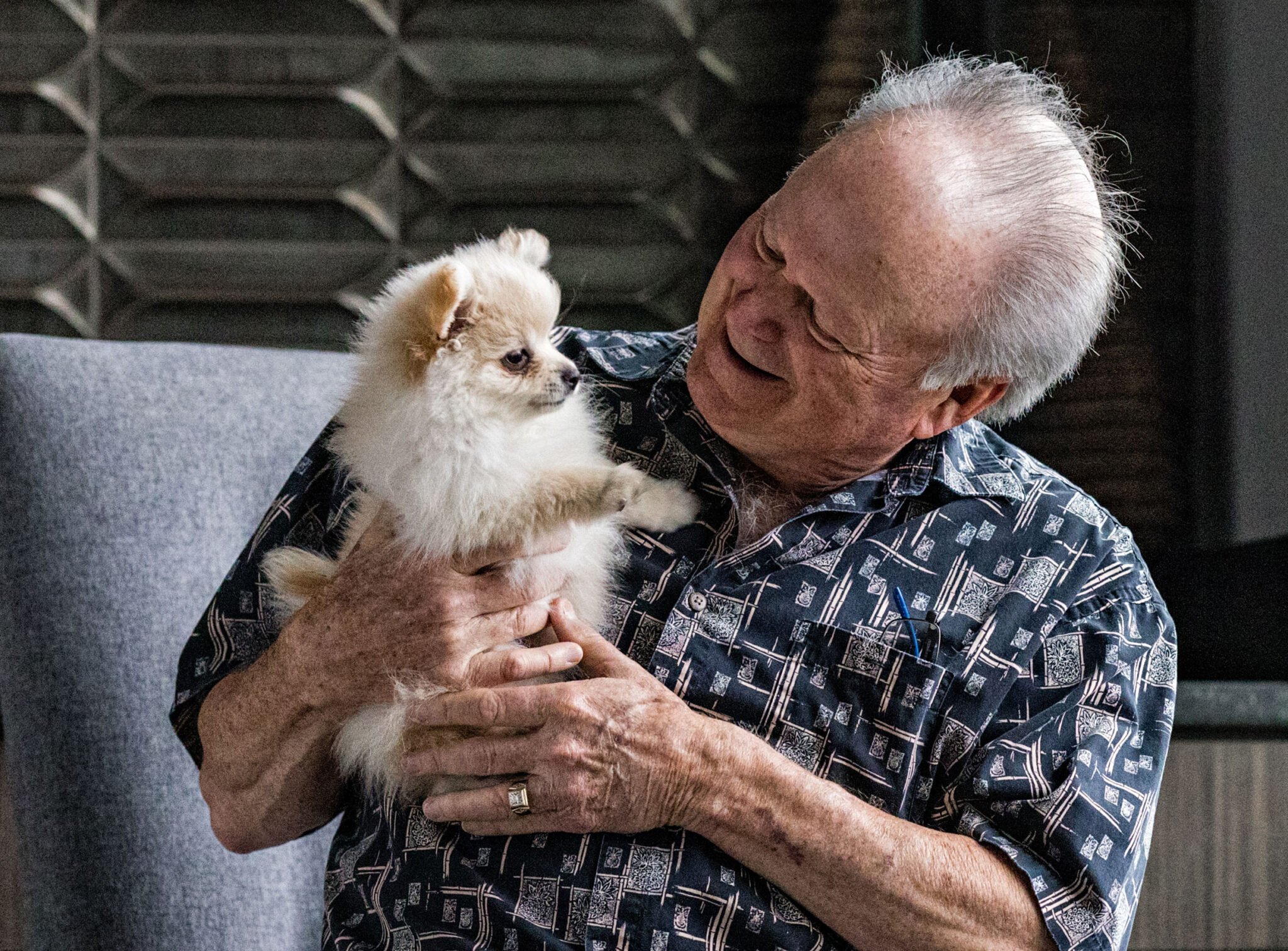 A senior man holds and smiles at a small white and tan colored dog