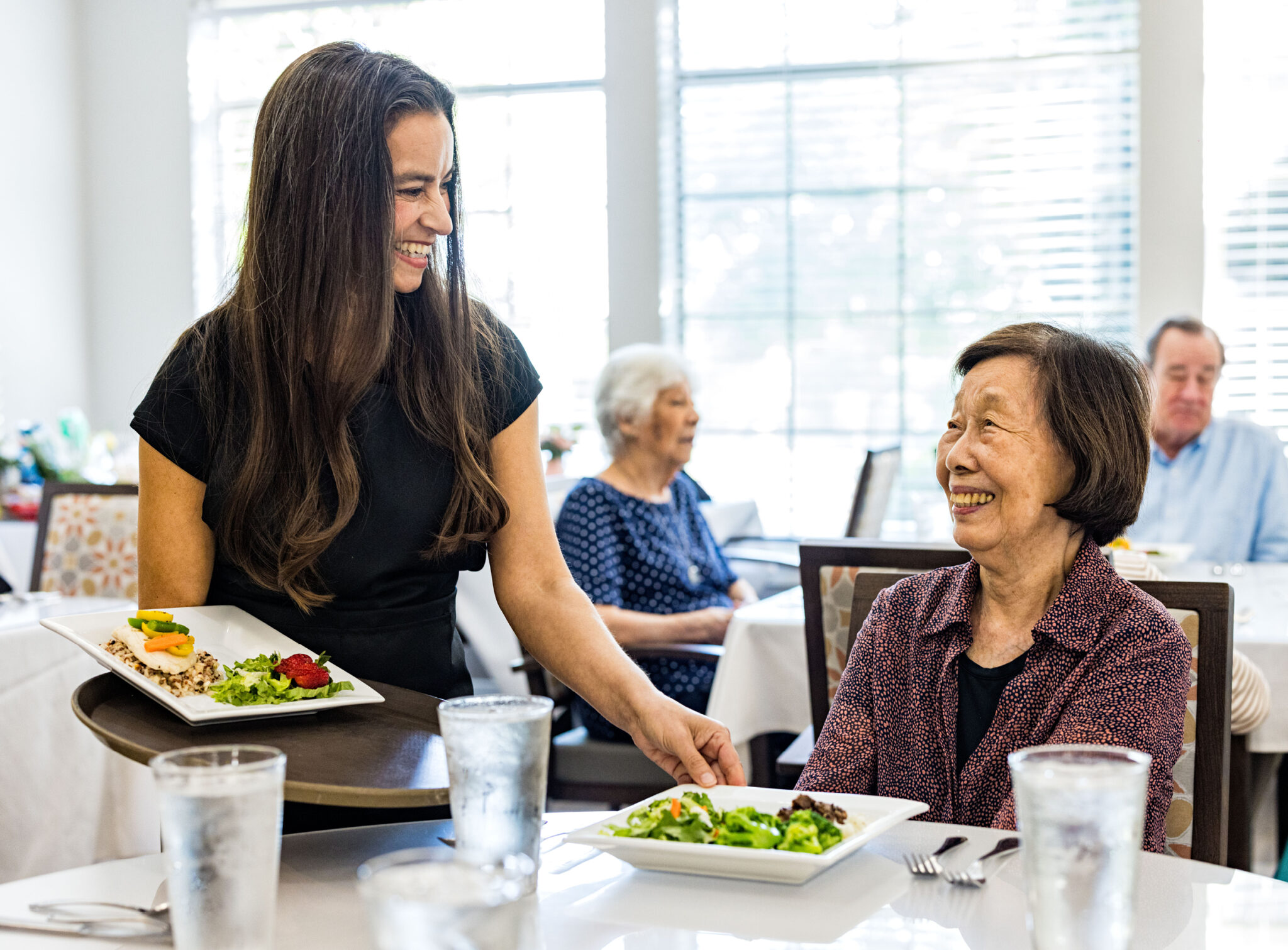 An older adult woman at a senior living community smiles while receiving her salad from a female caregiver