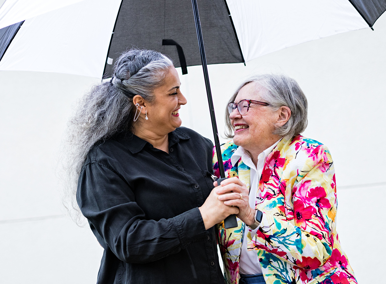 Senior female resident and an older adult female caregiver stand and smile while under an umbrella