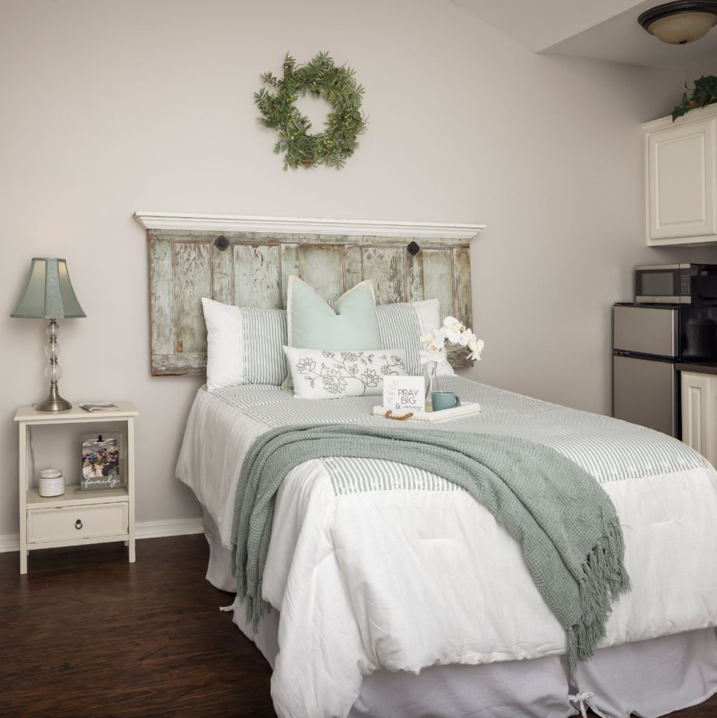 Apartment bedroom includes a full size bed, side table with a lamp and a kitchenette with a mini fridge and microwave