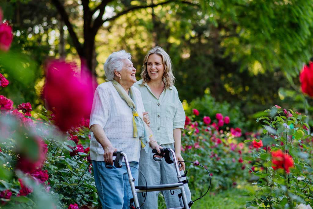 Elderly woman with walker and younger woman walk through a flower garden together