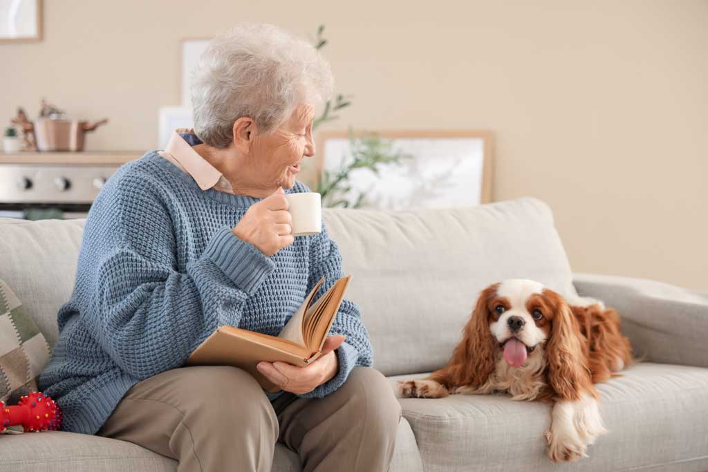 Elderly woman holding a book and tea while sitting on couch with dog